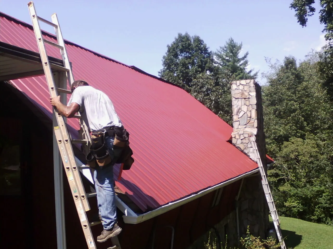 Roofer on a ladder beside a red metal roof and stone chimney — Soffit & Fascia Repair by Roofing Brothers in Darrington